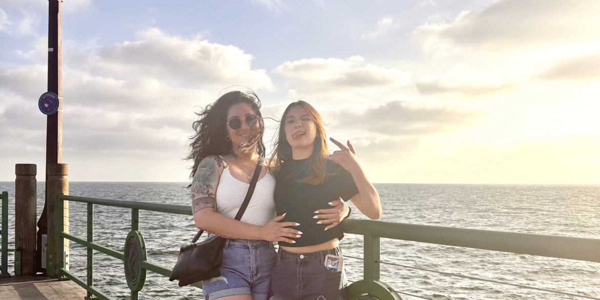 Madre migrante junto a su hija disfrutando un atardecer frente al mar, símbolo de unión y resiliencia familiar pese a la distancia.