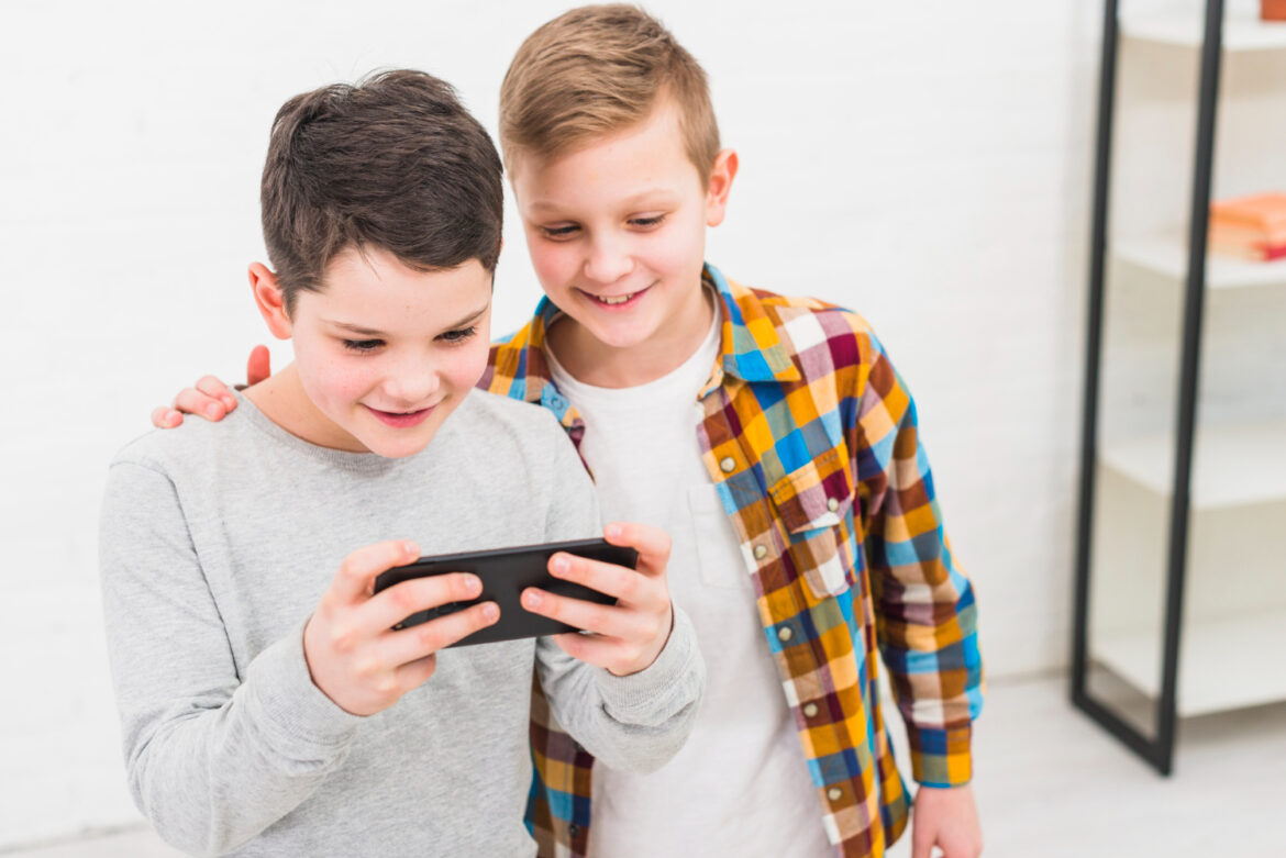 Dos niños sonríen mientras juegan juntos en un teléfono móvil, concentrados en la pantalla, en un entorno interior luminoso.