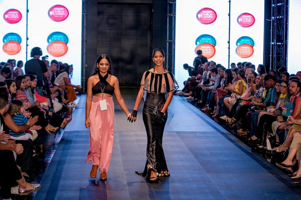 Diseñadora de modas nicaragüense acompaña a una modelo sobre la pasarela durante un desfile de moda, mientras los asistentes observan la presentación desde ambos lados del escenario.