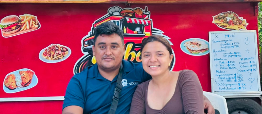 Una pareja sonriente sentada frente a un carrito de comida rápida de color rojo. El hombre viste una playera tipo polo azul y la mujer una blusa marrón, ambos posando para una foto profesional con iluminación mejorada y un fondo neutro y limpio.